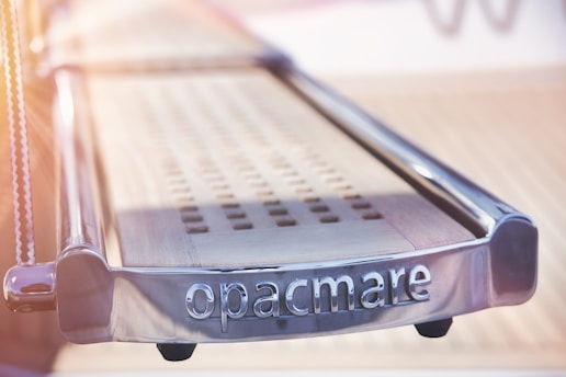 A polished metal boarding ladder with the word 'opacmare' engraved on it. The surface is shiny with a combination of metal and wooden elements, catching some natural light with a blurred background.