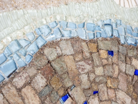 An intricate mosaic pattern composed of various small, closely arranged tiles. The upper section features light blue and clear glass tiles, smoothly transitioning into earth-toned stones below. Small, scattered pieces of bright blue glass add a striking contrast.