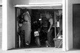 A monochrome image of a man standing in a cluttered garage, surrounded by various items like a surfboard, a motorcycle, and storage shelves filled with boxes. The man, with short, light-colored hair and tattoos, appears to be organizing or looking for something amidst the items.