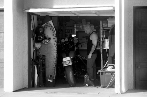 A monochrome image of a man standing in a cluttered garage, surrounded by various items like a surfboard, a motorcycle, and storage shelves filled with boxes. The man, with short, light-colored hair and tattoos, appears to be organizing or looking for something amidst the items.