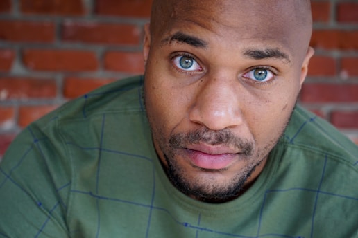 A person with a bald head is looking directly into the camera against a backdrop of red brick. Their eyes are a striking blue, and they are wearing a green shirt with a subtle check pattern.