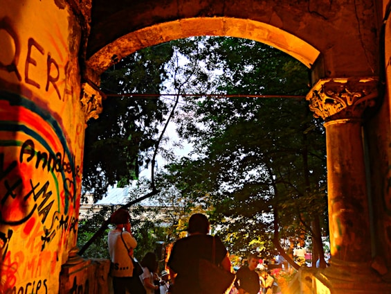 A historic stone archway with detailed carvings frames a view of a park or wooded area. The walls are covered with colorful graffiti art, featuring vibrant rainbow hues and assorted writing. People, including a person photographing, are gathered beneath the arch, creating a sense of community and liveliness.