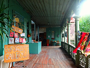 A veranda featuring rustic wooden floors and green-painted walls and ceilings, adorned with colorful art pieces and a welcoming sign. Several paintings are displayed on an easel with a 'Welcome' sign. Lush greenery is visible on the right. The area has an eclectic and artistic atmosphere, with fabric draped along the railing.