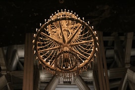 A large, ornate chandelier with multiple layers of intricate designs and numerous small lights that emit a warm glow. The lighting fixture is set against a darker background, possibly the ceiling of a grand hall or cathedral, emphasizing its elaborate craftsmanship.