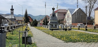 A well-maintained, cobblestone pathway leads through a neatly arranged garden flanked by yellow dandelions. A monument with a statue on top is centrally located. Residential and municipal buildings are visible in the background, along with mountains under a partly cloudy sky.