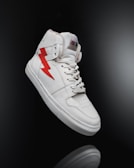 A white high-top sneaker prominently displayed against a dark background. The shoe features a distinctive red lightning bolt design on the side and is styled with white laces. The overall look is clean and modern, highlighting the shoe's sleek design and details.