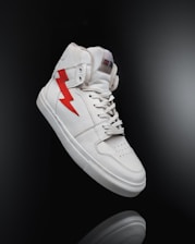 A white high-top sneaker prominently displayed against a dark background. The shoe features a distinctive red lightning bolt design on the side and is styled with white laces. The overall look is clean and modern, highlighting the shoe's sleek design and details.