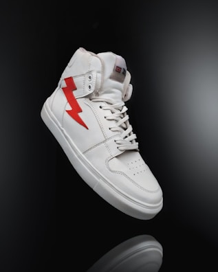 A white high-top sneaker prominently displayed against a dark background. The shoe features a distinctive red lightning bolt design on the side and is styled with white laces. The overall look is clean and modern, highlighting the shoe's sleek design and details.