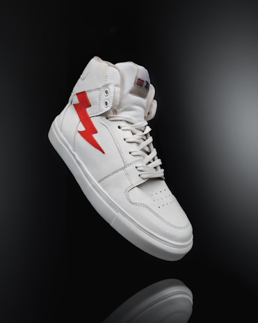 A white high-top sneaker prominently displayed against a dark background. The shoe features a distinctive red lightning bolt design on the side and is styled with white laces. The overall look is clean and modern, highlighting the shoe's sleek design and details.
