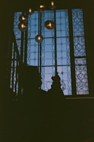 A dimly lit interior scene featuring a large stained glass window. The window has intricate geometric and floral patterns allowing a gentle blue hue to filter through. Several spherical pendant lights hang in front of the window, each glowing softly. Silhouettes, possibly of people, are seen in front of the window, adding to the mysterious ambiance.