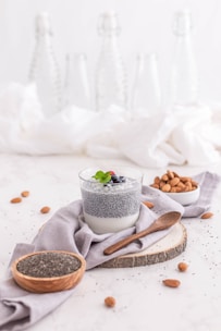 A glass of chia seed pudding topped with berries and mint is placed on a wooden slice with a wooden spoon nearby. Surrounding the main dish are scattered almonds, a small bowl filled with almonds, and a bowl containing chia seeds. The background includes blurred glass bottles and soft white fabric, contributing to a minimalist and clean presentation.