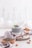 A glass filled with chia pudding placed on a wooden slab is garnished with mint leaves and berries. The setting includes a wooden spoon, scattered almonds, and a small bowl of chia seeds. A soft gray cloth drapes around the items, with a minimalist background featuring blurred bottles.