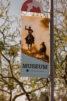 A banner hangs from a pole, displaying an image of a cowboy riding a horse with another horse running beside them. The cowboy is holding a lasso in the air, and the scene is set against a backdrop of dust and warm sunlight. The banner reads 'MUSEUM Smithsonian Affiliate' in bold letters. The surrounding area includes branches of trees with sparse leaves.
