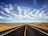 An open road stretches endlessly into the horizon under a vast, clear blue sky. Whispy clouds decorate the sky, creating a dynamic and eye-catching pattern. On either side of the road, dry grasslands extend outwards, with utility poles lining the roadside.