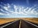 An open road stretches endlessly into the horizon under a vast, clear blue sky. Whispy clouds decorate the sky, creating a dynamic and eye-catching pattern. On either side of the road, dry grasslands extend outwards, with utility poles lining the roadside.