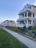 A row of large, multi-story houses with wrap-around balconies and manicured gardens. The homes are situated along a sidewalk and lush green lawns, under a clear blue sky. The scene is peaceful and well-maintained, reflecting a suburban or coastal neighborhood.