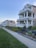 A row of large, multi-story houses with wrap-around balconies and manicured gardens. The homes are situated along a sidewalk and lush green lawns, under a clear blue sky. The scene is peaceful and well-maintained, reflecting a suburban or coastal neighborhood.