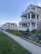 A row of large, multi-story houses with wrap-around balconies and manicured gardens. The homes are situated along a sidewalk and lush green lawns, under a clear blue sky. The scene is peaceful and well-maintained, reflecting a suburban or coastal neighborhood.