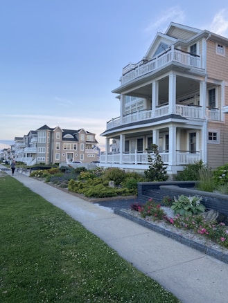A row of large, multi-story houses with wrap-around balconies and manicured gardens. The homes are situated along a sidewalk and lush green lawns, under a clear blue sky. The scene is peaceful and well-maintained, reflecting a suburban or coastal neighborhood.