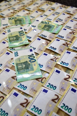 A spread of numerous 100 and 200 Euro banknotes scattered over a flat surface. The banknotes are new and crisp, with blue and yellow colors predominating. The arrangement appears somewhat disorganized, with some bills overlapping each other.