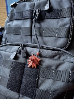 A close-up view of a black tactical backpack featuring multiple straps and Velcro patches. Attached to the backpack is a small, red pendant in the shape of a stylized wolf head.