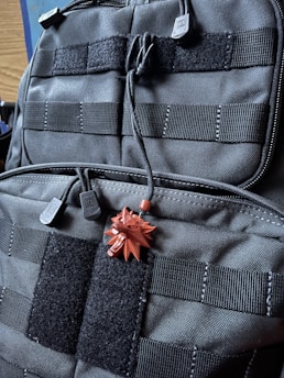 A close-up view of a black tactical backpack featuring multiple straps and Velcro patches. Attached to the backpack is a small, red pendant in the shape of a stylized wolf head.