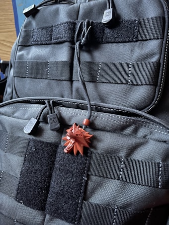 A close-up view of a black tactical backpack featuring multiple straps and Velcro patches. Attached to the backpack is a small, red pendant in the shape of a stylized wolf head.