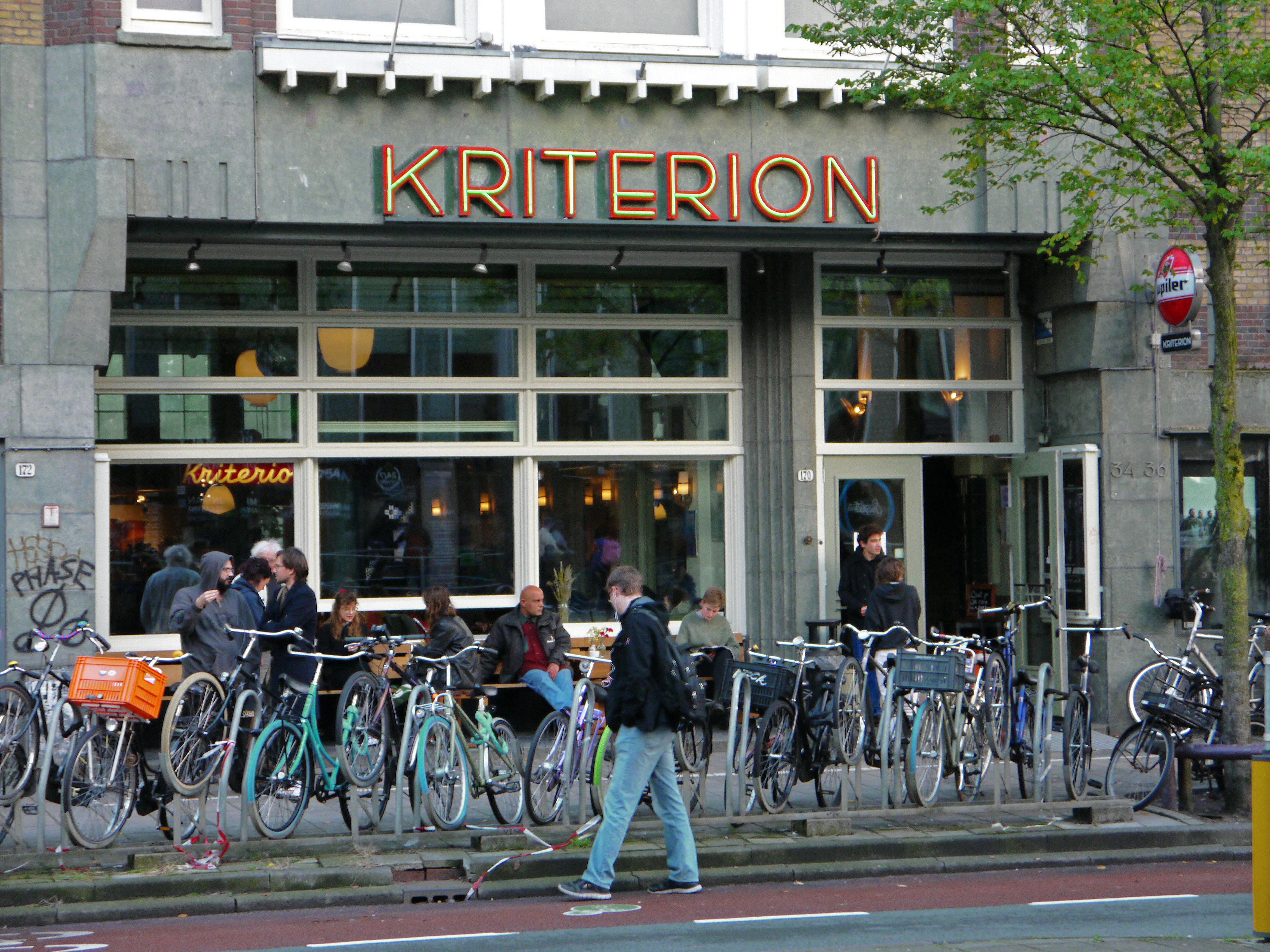 Photo of a building facade of Kriterion film theater and pub with young city people in front of the reflecting windows. The design of the gray facade is made in stone architecture of c. 1930; free street photography of Amsterdam city - Fons Heijnsbroek, The Netherlands October 2021
