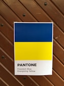 A poster displaying two horizontal color bands; the top is a bright blue labeled 'Freedom Blue' and the bottom is a vivid yellow labeled 'Energizing Yellow'. The poster features the word 'Pantone' printed below the colors. The background consists of a wooden panel with visible screws.