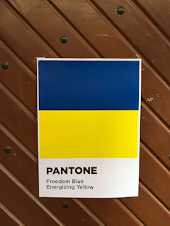 A poster displaying two horizontal color bands; the top is a bright blue labeled 'Freedom Blue' and the bottom is a vivid yellow labeled 'Energizing Yellow'. The poster features the word 'Pantone' printed below the colors. The background consists of a wooden panel with visible screws.