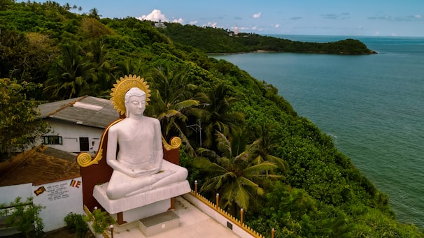 A large white statue of a seated Buddha with a golden halo is situated on a platform overlooking a lush, green, forested hill beside a blue ocean. A building with a brown-tiled roof is nearby, partially obscured by the dense surrounding vegetation. The sky is clear with a few scattered clouds.