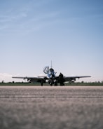 A small aircraft is parked on a runway with an overcast sky above. A solitary person stands near the aircraft, possibly engaged in a pre-flight inspection or maintenance task.