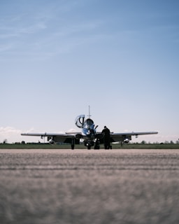 A small aircraft is parked on a runway with an overcast sky above. A solitary person stands near the aircraft, possibly engaged in a pre-flight inspection or maintenance task.