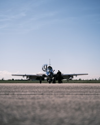 A small aircraft is parked on a runway with an overcast sky above. A solitary person stands near the aircraft, possibly engaged in a pre-flight inspection or maintenance task.