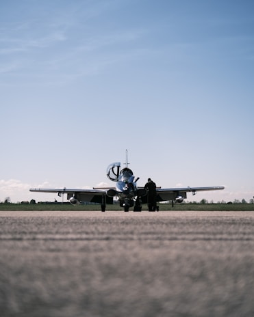 A small aircraft is parked on a runway with an overcast sky above. A solitary person stands near the aircraft, possibly engaged in a pre-flight inspection or maintenance task.