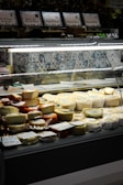 A display case in a store holds a variety of cheeses, ranging in color from white to dark orange. Several types of packaged cheeses are stacked neatly, with visible labels and price tags on some of them. Behind the display, there is a tiled wall with decorative blue and white patterns.
