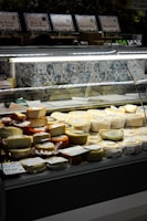 A display case in a store holds a variety of cheeses, ranging in color from white to dark orange. Several types of packaged cheeses are stacked neatly, with visible labels and price tags on some of them. Behind the display, there is a tiled wall with decorative blue and white patterns.