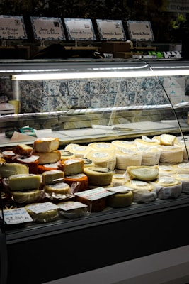 A display case in a store holds a variety of cheeses, ranging in color from white to dark orange. Several types of packaged cheeses are stacked neatly, with visible labels and price tags on some of them. Behind the display, there is a tiled wall with decorative blue and white patterns.