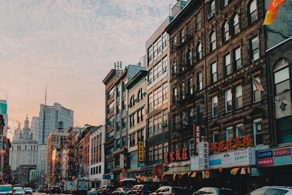 A bustling urban street scene featuring a row of older brick and modern glass buildings lined with vibrant signage in Chinese characters. The street is filled with vehicles and a few pedestrians, suggesting a lively atmosphere. The sky above is a mix of soft colors from the setting or rising sun, adding warmth to the scene.