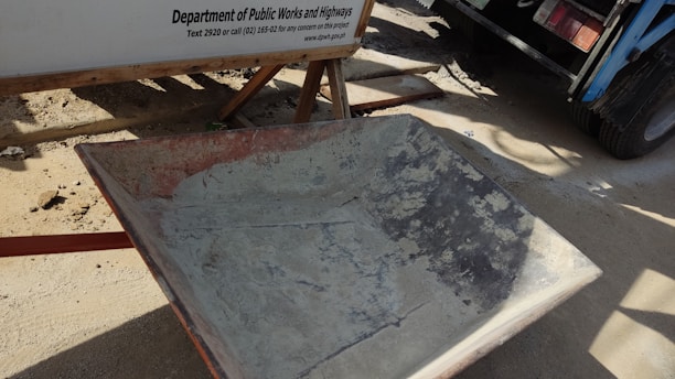 A weathered wheelbarrow with visible signs of rust stands on a dirt surface next to a construction sign from the Department of Public Works and Highways. In the background, part of a truck and its wheel are partially visible, casting shadows on the ground.