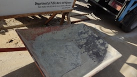 A weathered wheelbarrow with visible signs of rust stands on a dirt surface next to a construction sign from the Department of Public Works and Highways. In the background, part of a truck and its wheel are partially visible, casting shadows on the ground.
