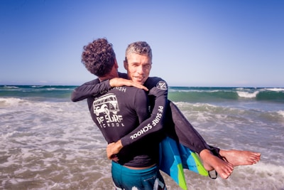 A man is standing in shallow water at the beach, carrying another person on his back. Both are wearing black rash guard shirts with a surf school logo and both holding surfboards. The ocean waves are visible in the background under a clear blue sky.