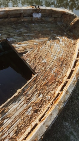 An old, weathered wooden boat with heavily damaged planks and exposed fibers. The boat is partially submerged in water with reflections of trees seen on the surface. The wood appears brittle and sun-bleached, indicating significant time spent exposed to the elements.