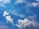 A clear blue sky is adorned with fluffy, white clouds scattered across the canvas. The clouds appear soft and billowy, creating gentle shapes against the vibrant azure background. Light wisps of clouds are seen blending with the deeper blues, suggesting a calm and peaceful atmosphere.