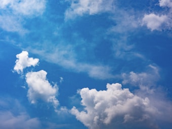 A clear blue sky is adorned with fluffy, white clouds scattered across the canvas. The clouds appear soft and billowy, creating gentle shapes against the vibrant azure background. Light wisps of clouds are seen blending with the deeper blues, suggesting a calm and peaceful atmosphere.