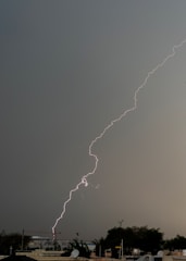 A bright streak of lightning cuts through a dark, overcast sky above an urban area. The ground is visible with buildings and trees below, suggesting a city landscape. The sky's heavy gray clouds contrast sharply with the bright, jagged lightning bolt.
