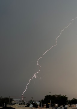 A bright streak of lightning cuts through a dark, overcast sky above an urban area. The ground is visible with buildings and trees below, suggesting a city landscape. The sky's heavy gray clouds contrast sharply with the bright, jagged lightning bolt.