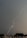 A bright streak of lightning cuts through a dark, overcast sky above an urban area. The ground is visible with buildings and trees below, suggesting a city landscape. The sky's heavy gray clouds contrast sharply with the bright, jagged lightning bolt.
