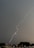A bright streak of lightning cuts through a dark, overcast sky above an urban area. The ground is visible with buildings and trees below, suggesting a city landscape. The sky's heavy gray clouds contrast sharply with the bright, jagged lightning bolt.