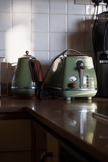 A kitchen countertop featuring a classic green kettle and toaster set. The countertop is made of a smooth, reflective surface. The kettle has a brown handle and a polished metal spout, while the toaster has round buttons and a dial. The background consists of white tiled walls and a few additional kitchen appliances partially visible.